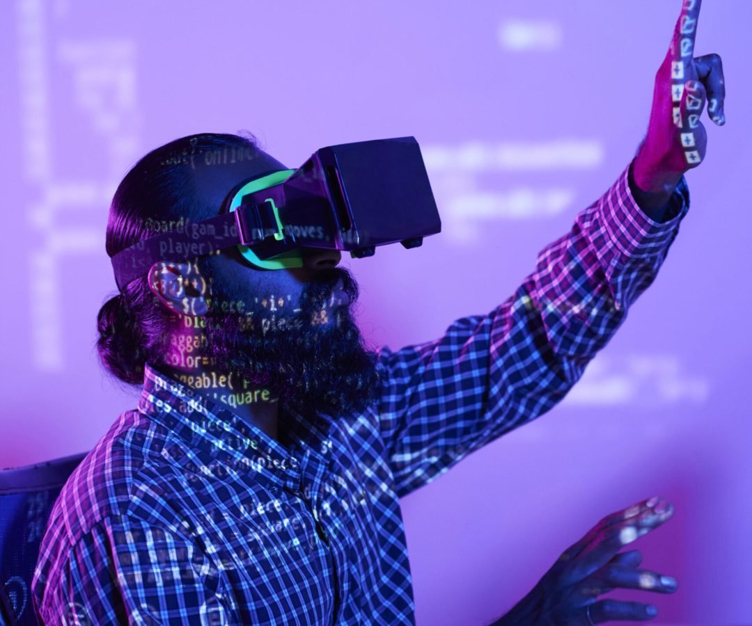 Man in vr headset touching virtual display while sitting in front of computer screen with coded data